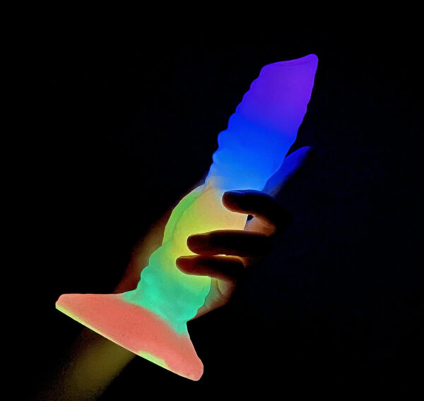 A hand holding a vibrant dildo against a dark background, showcasing its colorful design and glow.