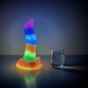 A vibrant rainbow-colored fantasy dildo sits beside a clear glass of water on a shiny surface.