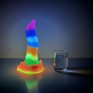 A vibrant rainbow-colored fantasy dildo sits beside a clear glass of water on a shiny surface.